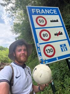 James Lewis standing next to a road sign marking the border of France, football at his feet, during his charity challenge for St Giles Hospice