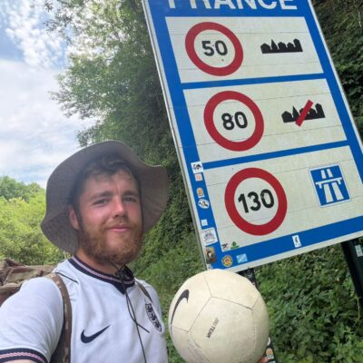 James Lewis standing next to a road sign marking the border of France, football at his feet, during his charity challenge for St Giles Hospice