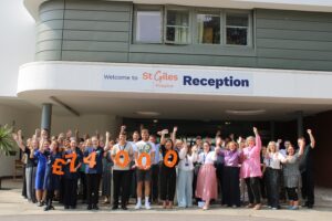 James Lewis surrounded by cheering St Giles Hospice staff holding orange numbers spelling £74,000 outside the hospice reception