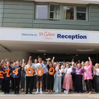 James Lewis surrounded by cheering St Giles Hospice staff holding orange numbers spelling £74,000 outside the hospice reception