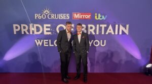 James Lewis and Sam Colclough from St Giles Hospice in suits and orange bow ties, standing in front of the Pride of Britain Awards backdrop on the red carpet.