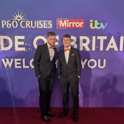 James Lewis and Sam Colclough from St Giles Hospice in suits and orange bow ties, standing in front of the Pride of Britain Awards backdrop on the red carpet.