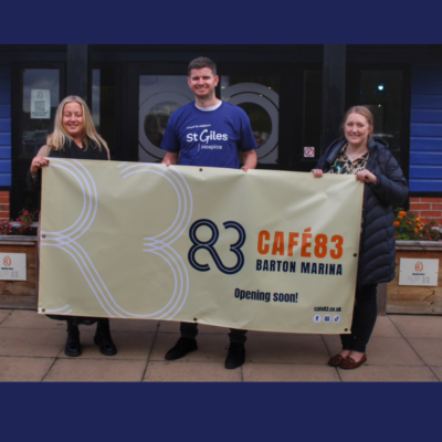 Three members of the St Giles Hospice team standing in a row outside Café 83 at Barton Marina, holding a Café 83 sign.